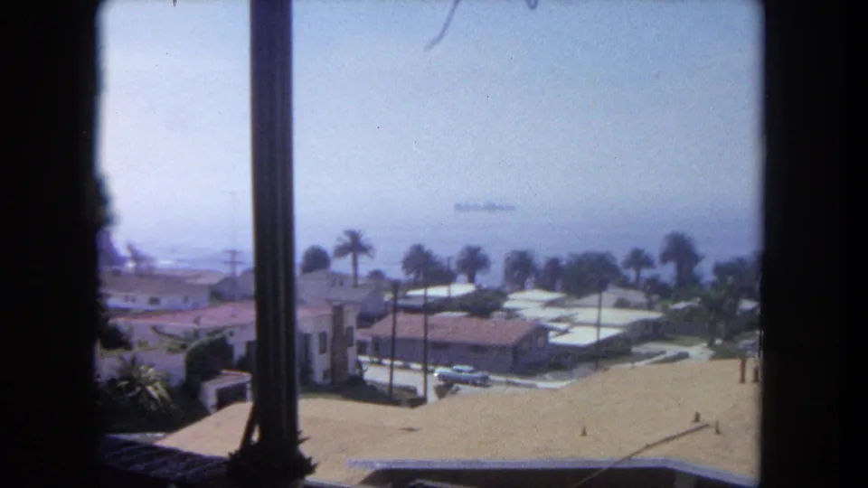 1962 Tijuana home-movie frame looking past a window post to coastal houses, palms, ocean haze, and a ship offshore.