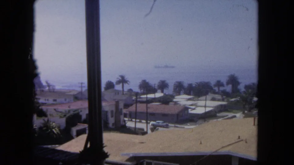 1962 Tijuana coastal neighborhood with a window edge in front, rooftops below, palms along the street, and a ship on the horizon.