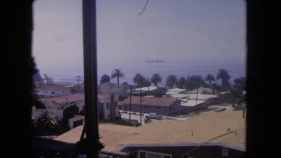 Final 1962 Tijuana frame shows a window-framed coastal neighborhood, palm trees, rooftops, parked cars, and a ship at sea.