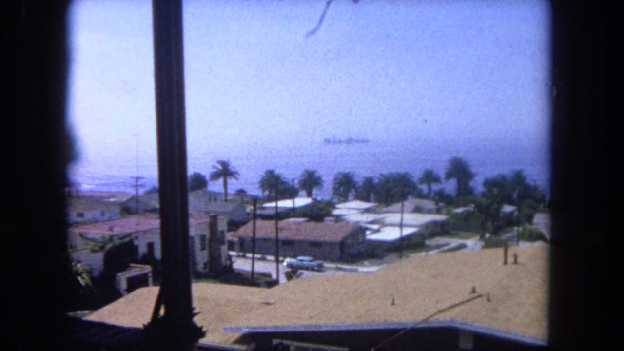 Elevated 1962 home-movie view near Tijuana showing beachside houses, palm trees, parked cars, ocean water, and a distant ship offshore.