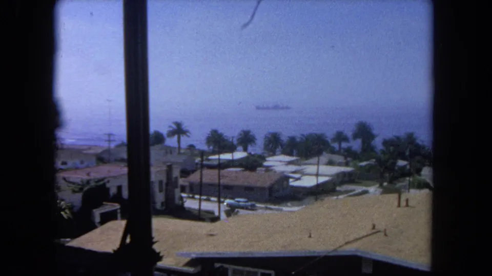 1962 8mm frame looking across a coastal residential area near Tijuana, with palms, low houses, sandy ground, ocean haze, and a ship offshore.