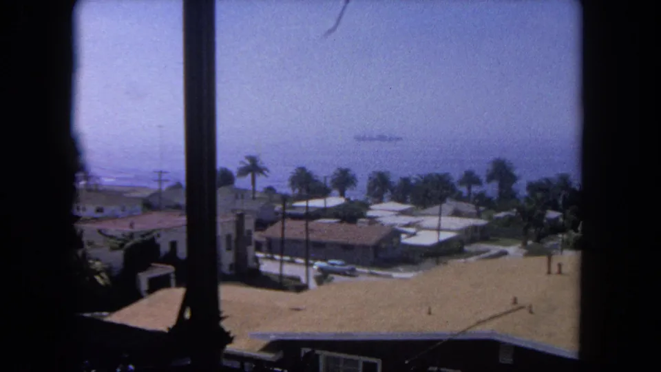 Sunlit 1962 home-movie view near Tijuana of beachside rooftops, palms, parked cars, and a faint ship on the water beyond the neighborhood.
