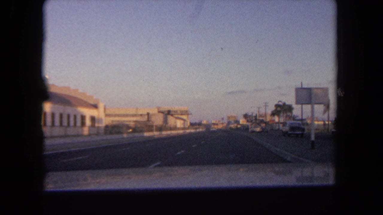 Roadway seen through a car window with low buildings, parked cars, signs, and palm trees at dusk.