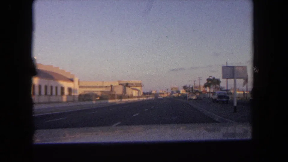 1962 Tijuana-area road view from a moving car, with low roadside buildings, signs, parked cars, and palm trees under fading light.