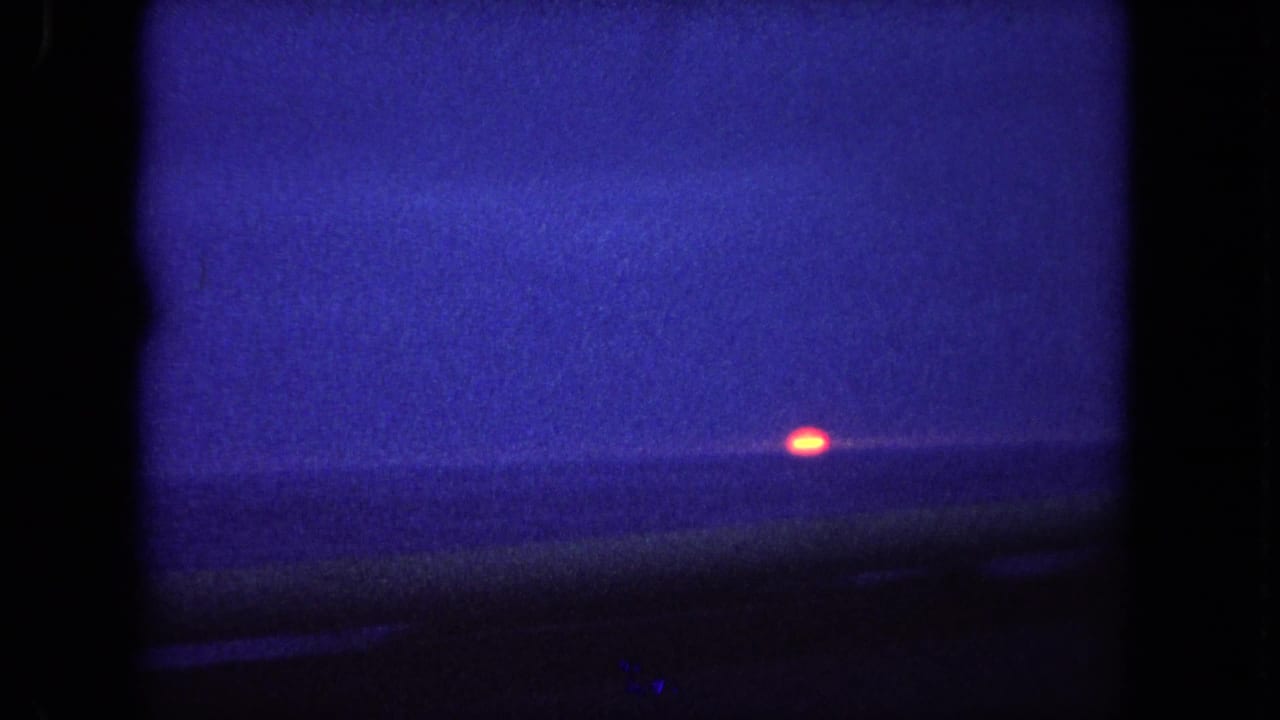 Blue-toned 8mm view of the ocean horizon near Tijuana, with a small orange sun framed by the dark edge of a car window.
