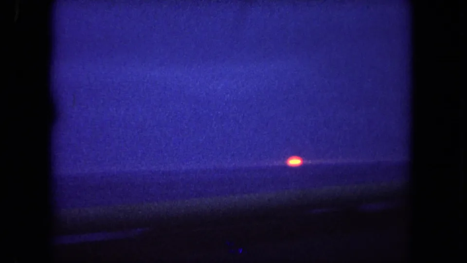 Blue-toned 8mm view of the ocean horizon near Tijuana, with a small orange sun framed by the dark edge of a car window.