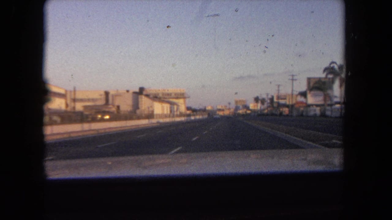 Forward view from a moving car on a multi-lane road at dusk with roadside buildings and palms.