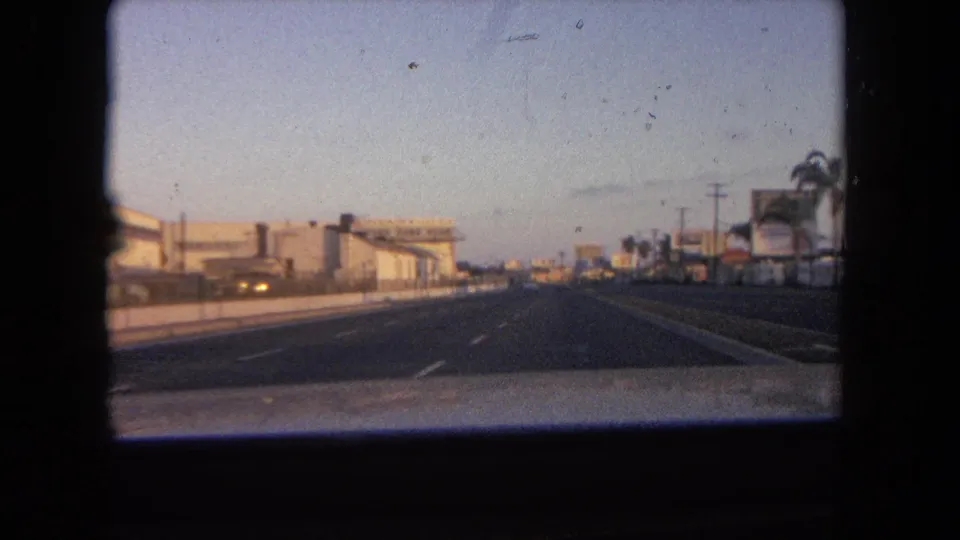 1962 Tijuana road view from inside a moving car, showing a wide roadway, low commercial buildings, palms, and fading sky.