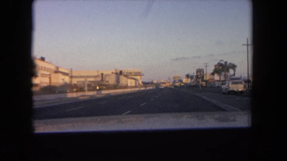 1962 Tijuana road scene from a moving vehicle, with low buildings, a roadside 45 sign, palms, and open lanes at dusk.