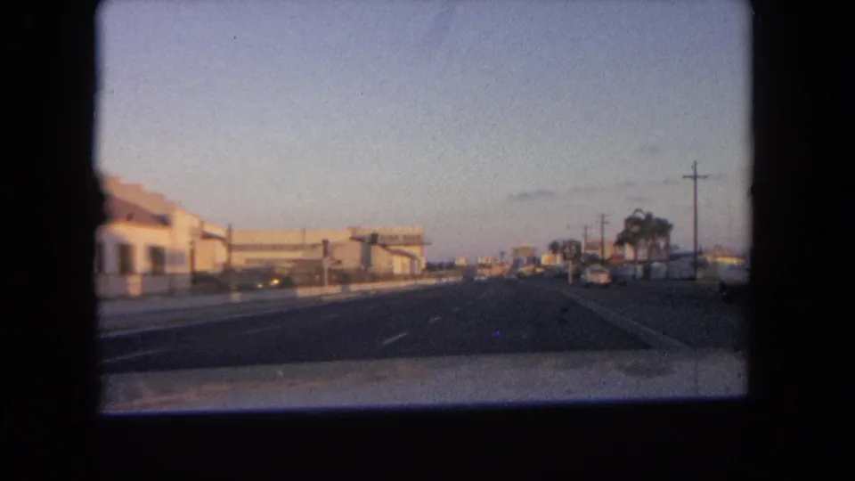 1962 Tijuana home-movie frame showing a car-level view down a broad roadway with commercial buildings and palms under fading light.