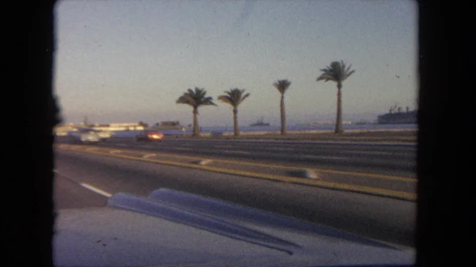 1962 Tijuana home-movie view from inside a car, looking across a divided road with palm trees, traffic, water, and a distant ship.