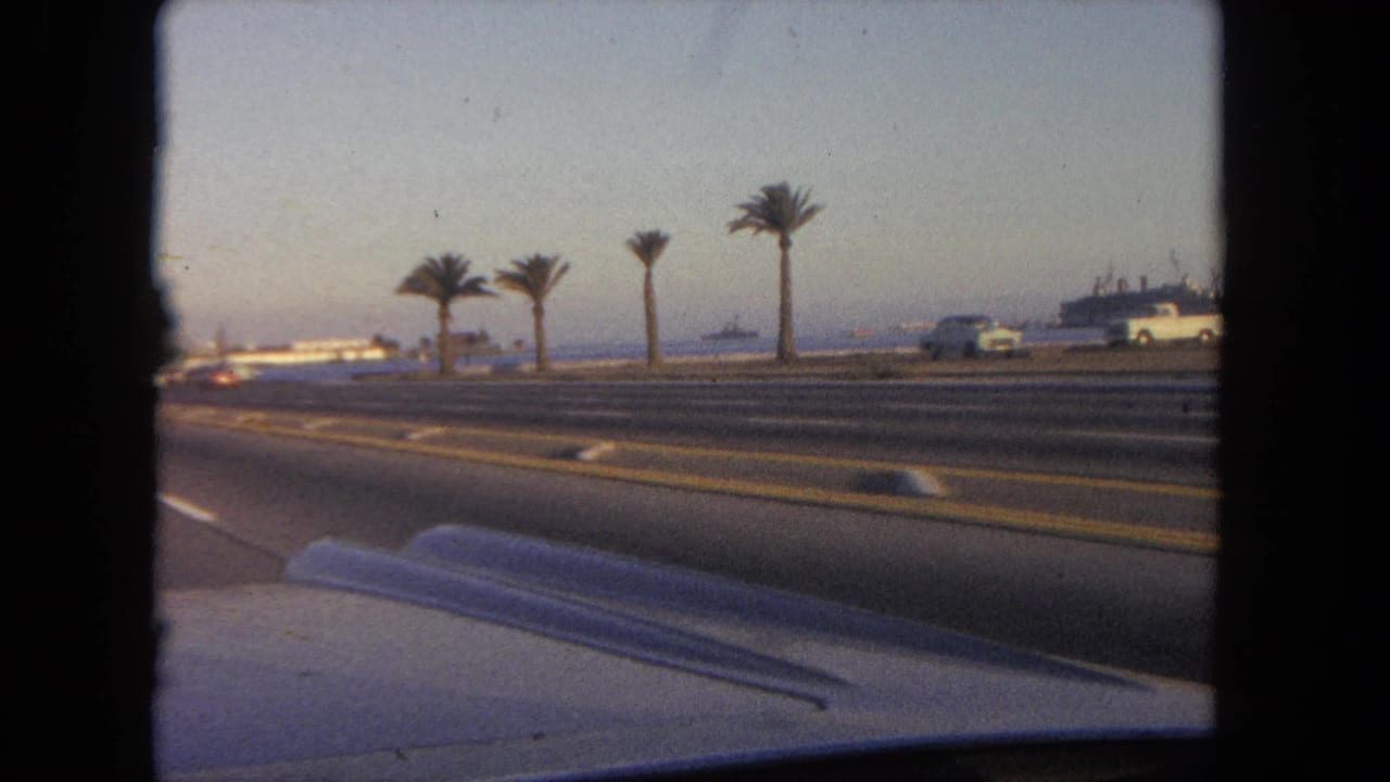 The in-car view continues along a Tijuana coastal road in 1962, with palms on the median, traffic, a shoreline, and a large vessel beyond.