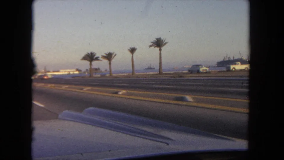 The in-car view continues along a Tijuana coastal road in 1962, with palms on the median, traffic, a shoreline, and a large vessel beyond.