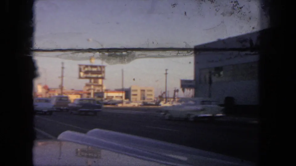 A 1962 Tijuana street view from inside a car shows traffic, low buildings, utility poles, and a roadside billboard, with heavy film wear visible.