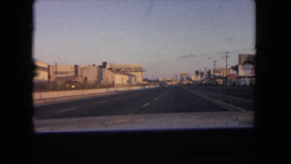 The car moves along a broad Tijuana roadway in 1962, passing low urban buildings, lane markings, utility poles, and palms under a clear sky.