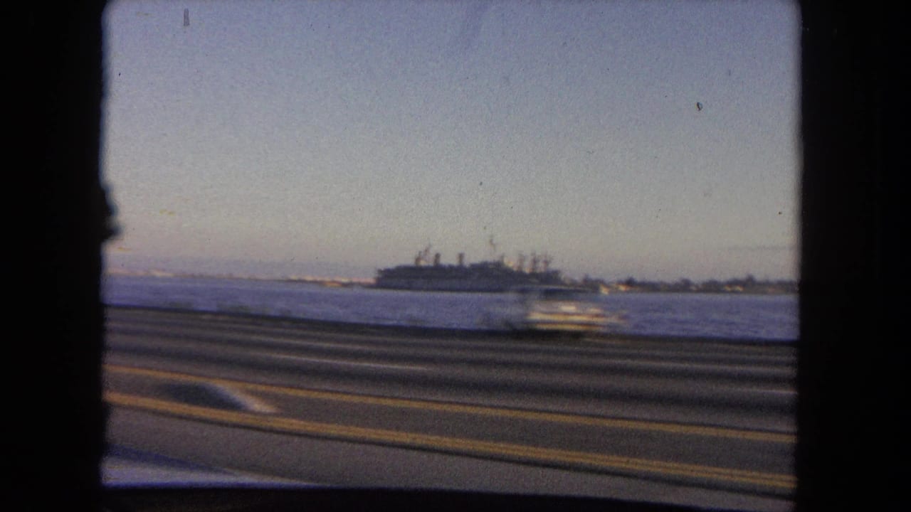 Coastal road seen from a moving car with a large ship offshore.
