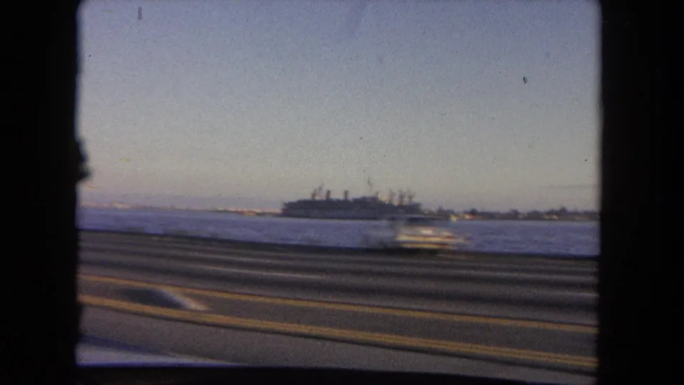 1962 Tijuana coastal drive view from a moving car, with highway lanes in front and a large ship visible on the water.