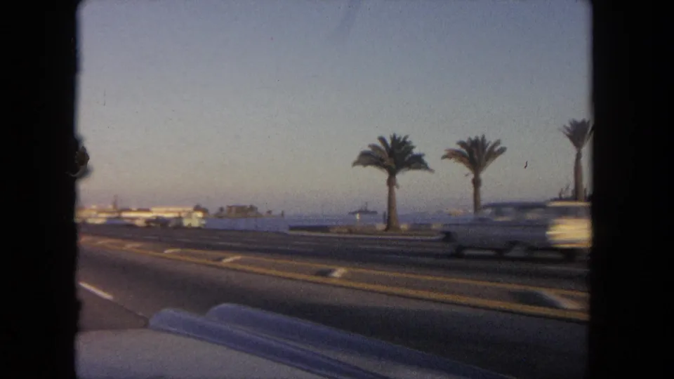 1962 Tijuana coastal drive scene with several palm trees, road lanes, passing cars, and the sea visible beyond the roadside.