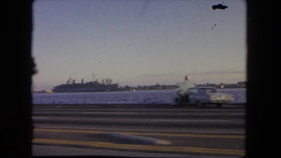 1962 Tijuana home-movie view from a moving vehicle, showing a docked ship across water and a car passing on the highway.