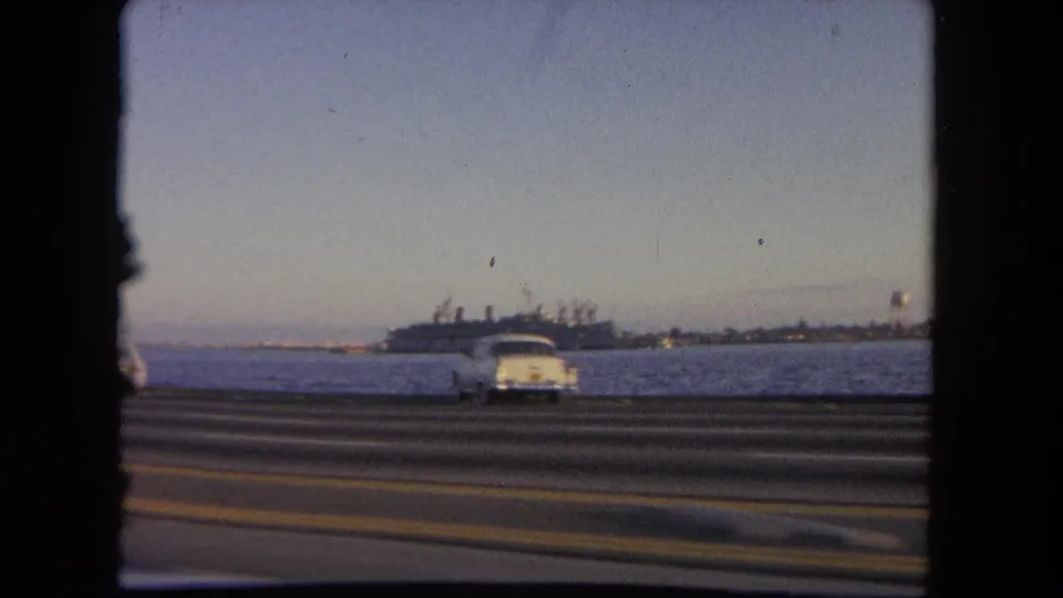 A white classic car passes along the highway in this 1962 Tijuana waterfront view, with water and a large ship beyond the road.