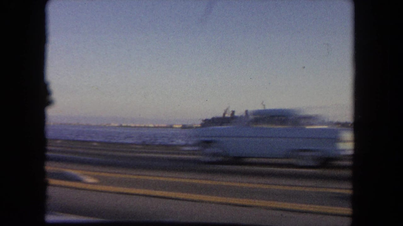 View from a moving car past a coastal road with a naval ship offshore.