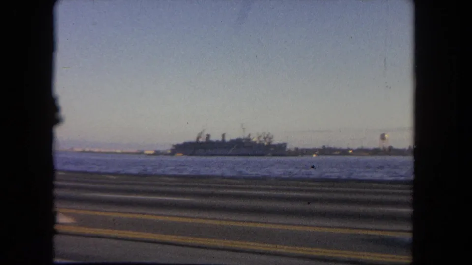 1962 Tijuana waterfront view from a moving vehicle, showing a road rail, sea, distant shoreline, and large naval vessel.