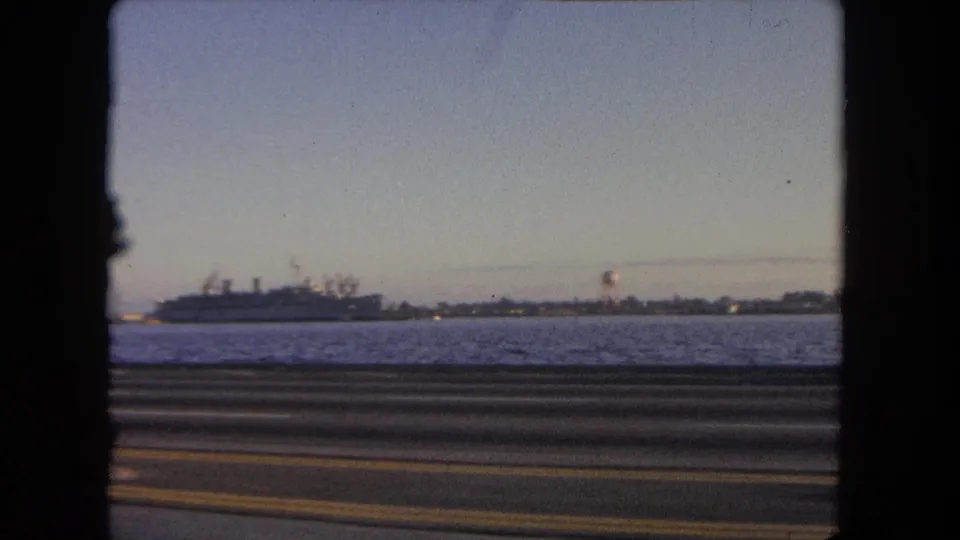 1962 Tijuana coastal driving shot with blurred roadway foreground, open water, shoreline, and large naval ship offshore.