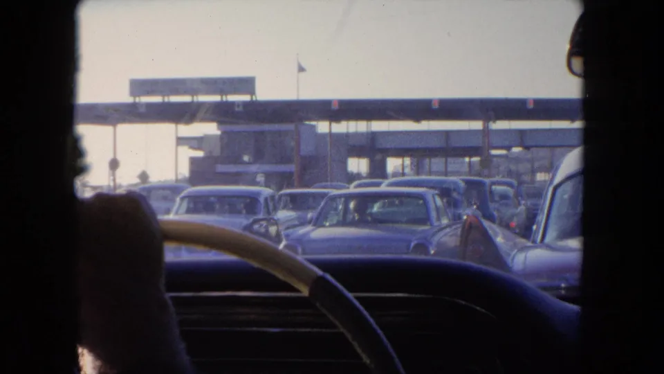 1962 Tijuana home-movie frame from inside a car, looking past a steering wheel toward mid-century traffic near the water.