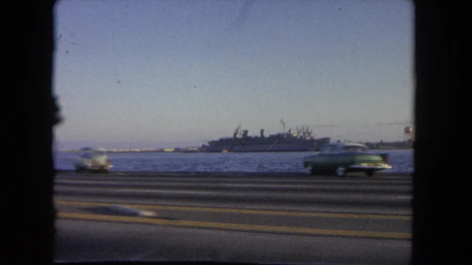 1962 Tijuana home-movie frame showing motion-blurred traffic along a waterfront road with a large ship offshore or docked beyond.