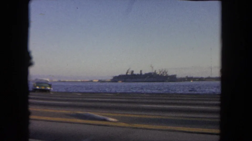 1962 Tijuana waterfront driving frame with a road in the foreground and a large ship across the water under a pale blue sky.