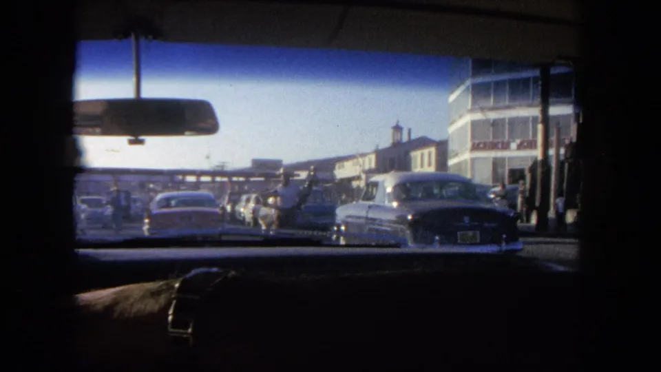 1962 Tijuana home-movie view from inside a car, looking through the windshield at vehicles queued near a toll plaza.