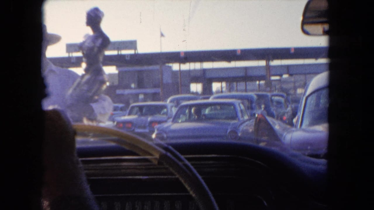 In-car 8mm view in Tijuana showing the steering wheel, dashboard, and lanes of mid-century cars approaching a toll area.