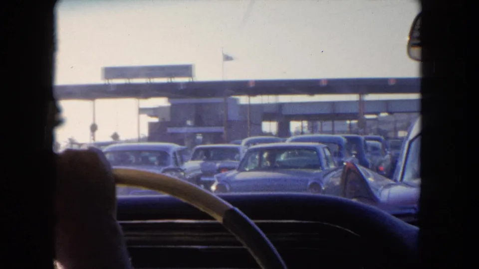 1962 Tijuana traffic seen over the dashboard, with multiple cars waiting beneath a toll-plaza canopy.
