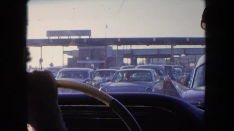 Final in-car view of a 1962 Tijuana toll approach, with traffic packed ahead and the windshield framing the plaza.