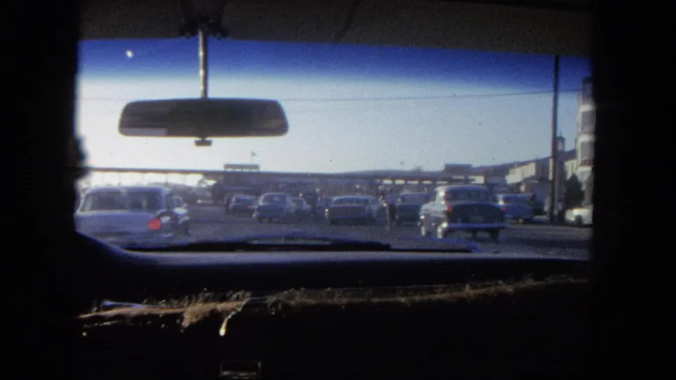 The car continues through traffic, showing 1960s-era vehicles, storefront edges, and the windshield framing.