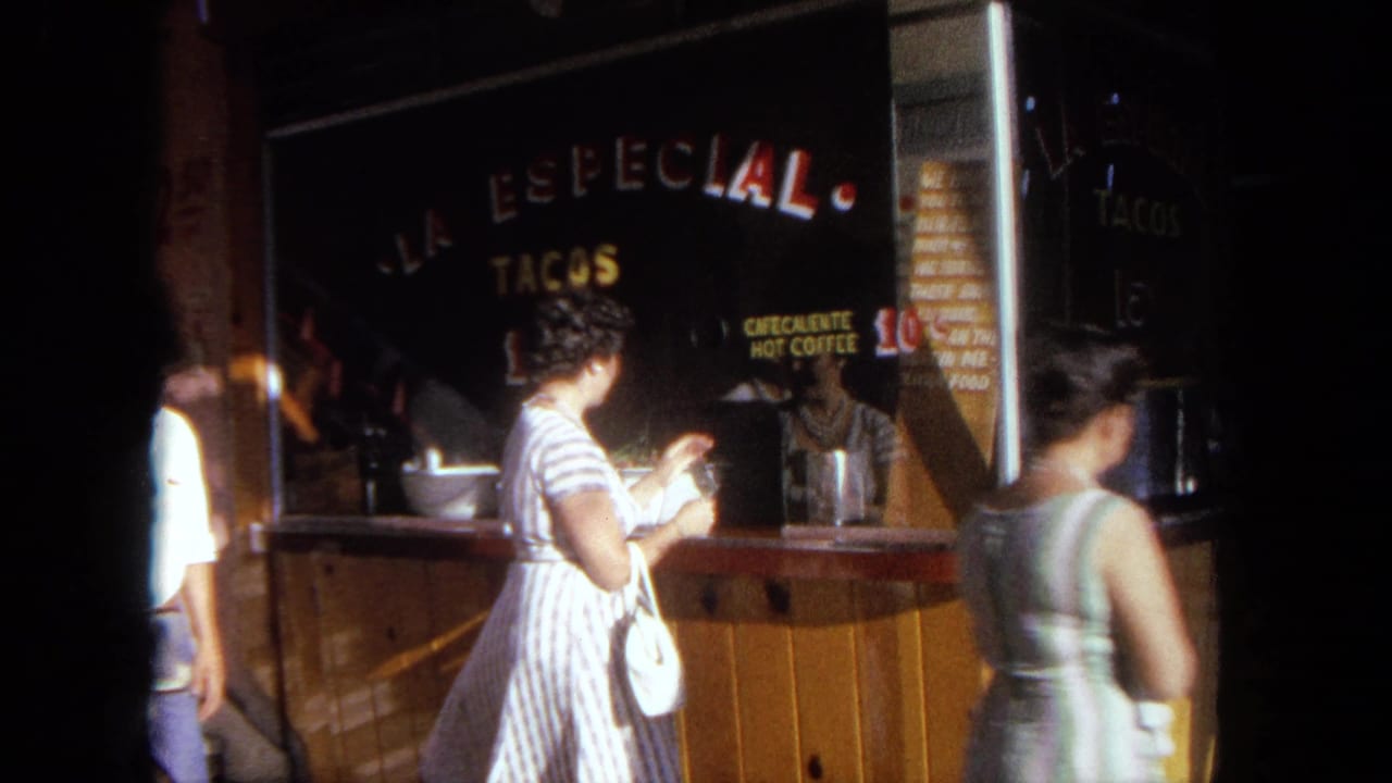 1962 Tijuana home-movie frame showing a woman with a white purse at a wooden taco stand with painted signs.