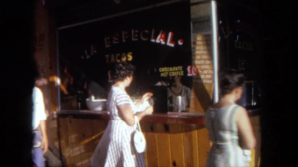 1962 Tijuana home-movie frame showing a woman with a white purse at a wooden taco stand with painted signs.