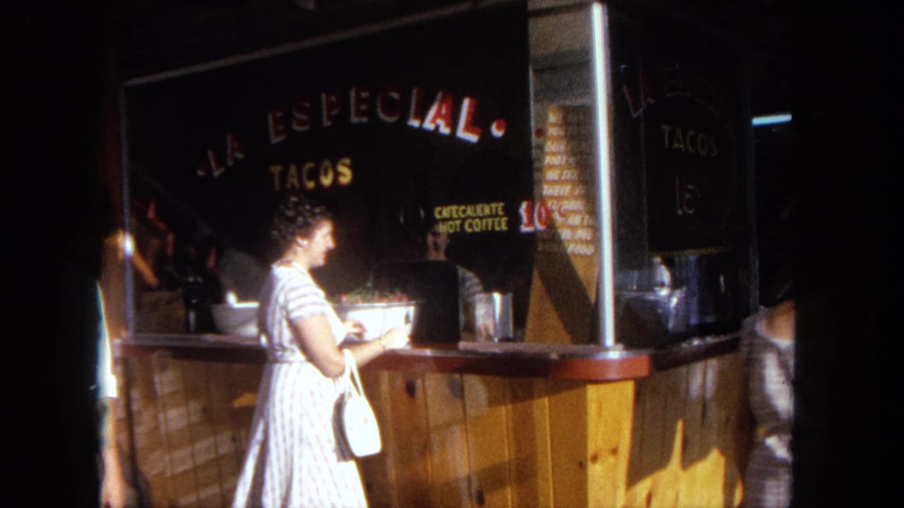 1962 Tijuana frame with a woman at a taco counter, readable La Especial signage, and a dim nighttime setting.
