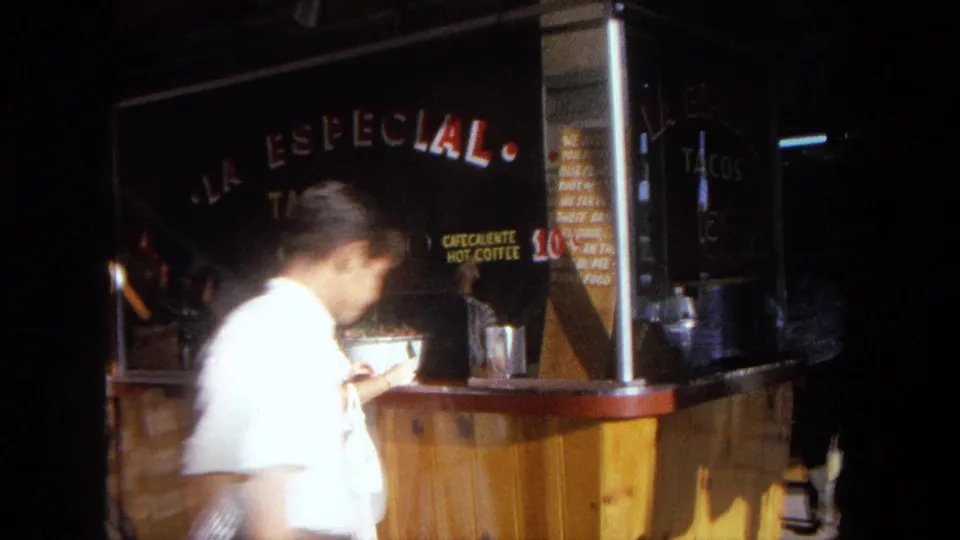 1962 Tijuana home-movie frame where a passerby briefly obscures the woman ordering at the taco stand.