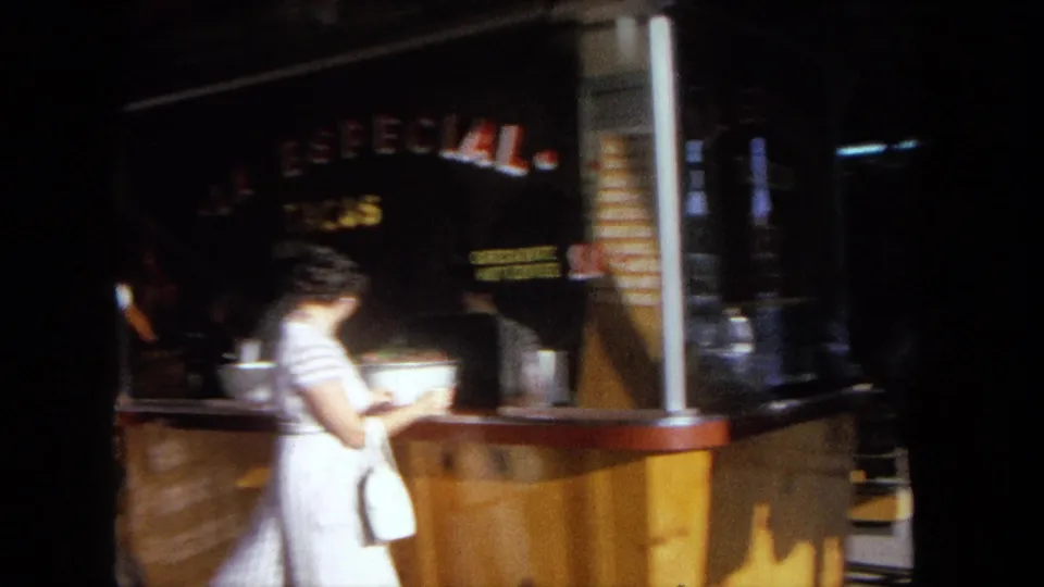 1962 Tijuana frame showing the woman beside the wooden taco stand as the handheld film catches night movement and blur.