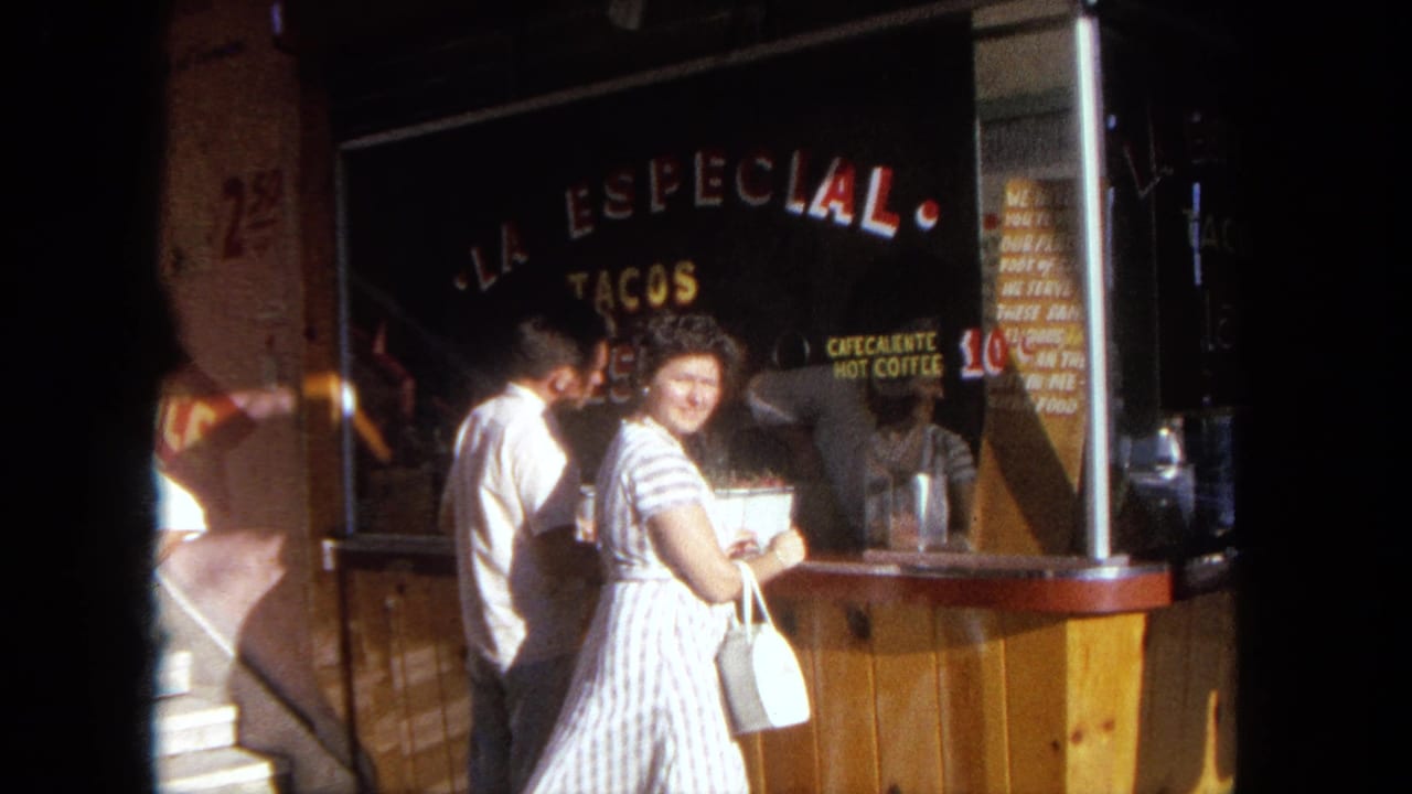 In 1962 Tijuana, a woman in a striped dress smiles toward the camera at an outdoor taco stand with men standing nearby.