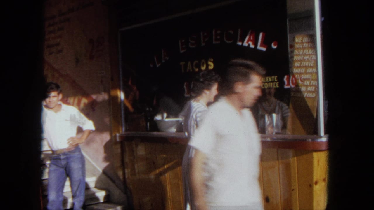 A passerby crosses the foreground as the woman and taco stand signage remain partly visible in this 1962 Tijuana home-movie frame.
