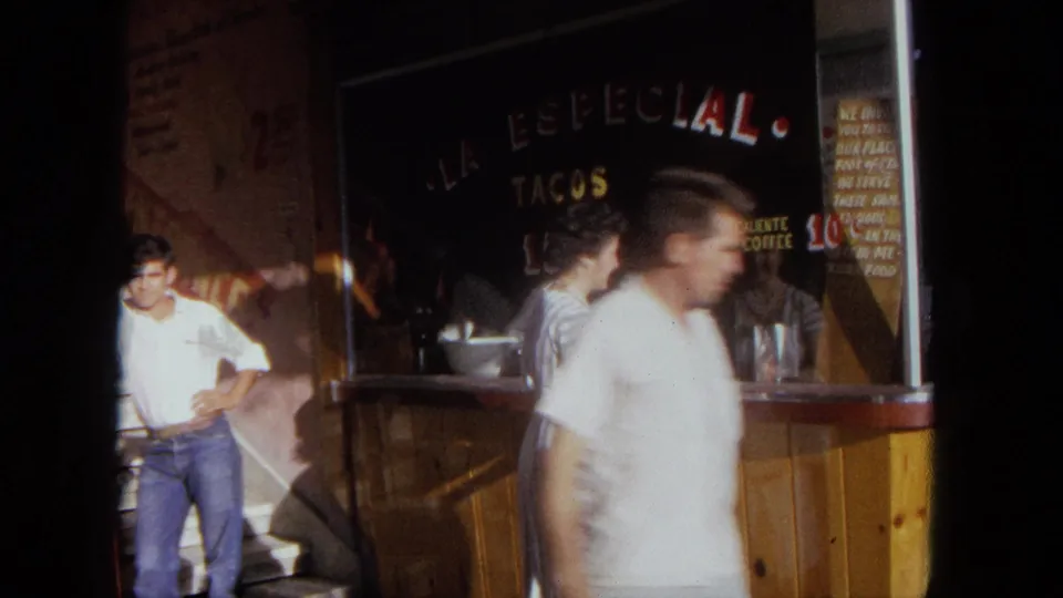 A passerby crosses the foreground as the woman and taco stand signage remain partly visible in this 1962 Tijuana home-movie frame.