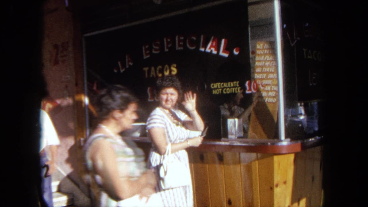 The woman waves toward the camera at La Especial Tacos in Tijuana while another pedestrian passes through the foreground.