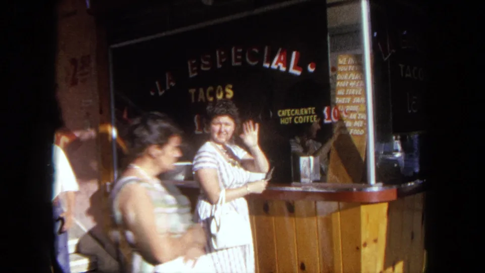 The woman waves toward the camera at La Especial Tacos in Tijuana while another pedestrian passes through the foreground.