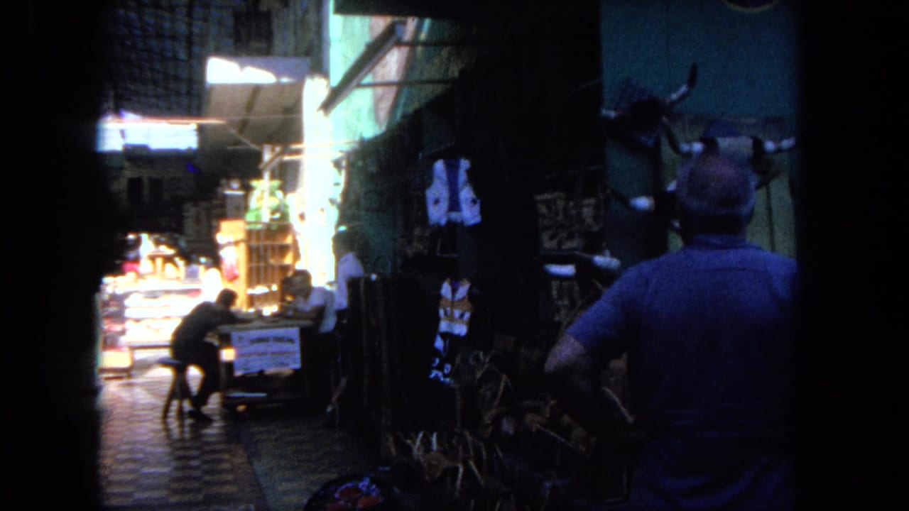 1962 Tijuana home-movie frame showing a man seen from behind in a narrow market alley with mounted horns, woven goods, and pedestrians.