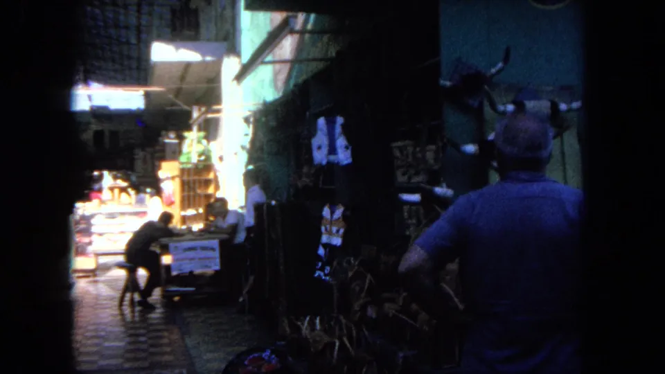 1962 Tijuana home-movie frame showing a man seen from behind in a narrow market alley with mounted horns, woven goods, and pedestrians.