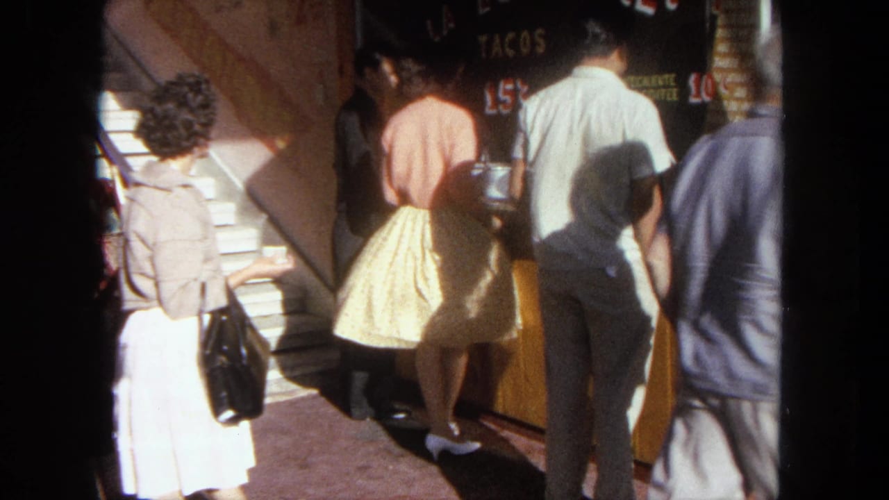 1962 Tijuana street-market frame showing pedestrians clustered by a taco stand, painted TACOS signage, yellow counter, and nearby stairs.