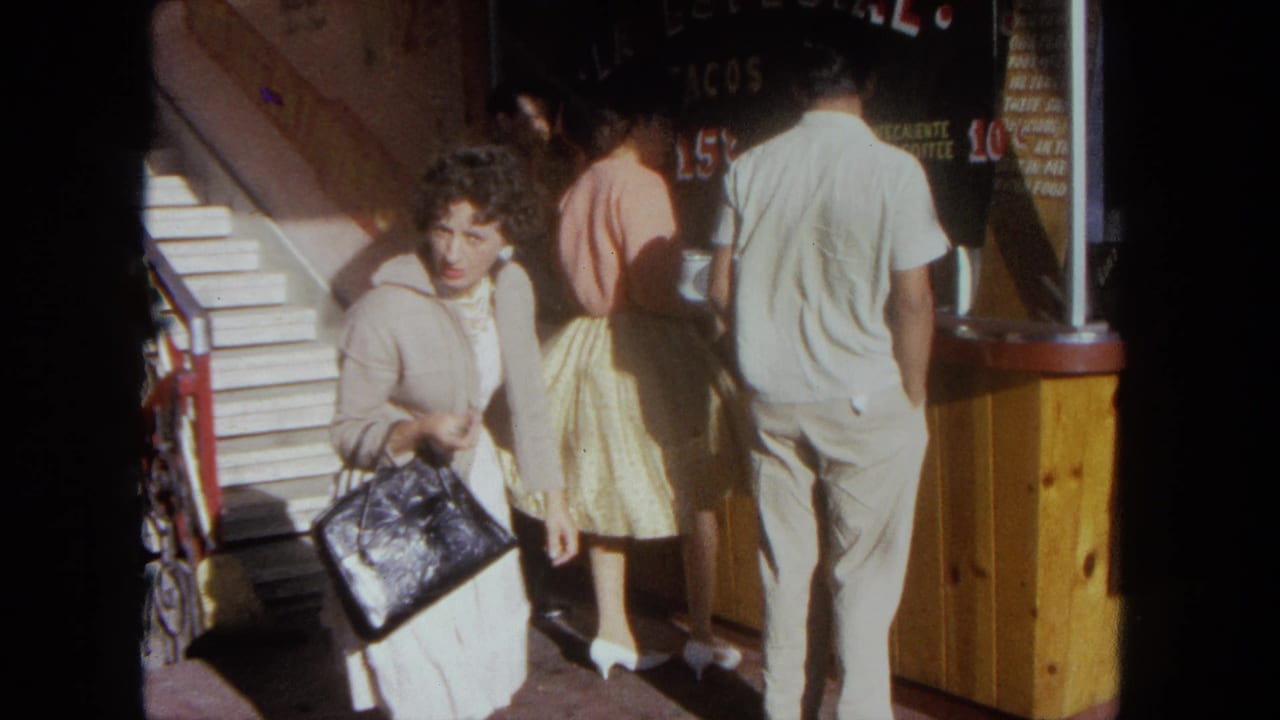 1962 Tijuana home-movie frame with a woman holding a handbag near a taco stand, pedestrians, painted signage, and sunlit steps.