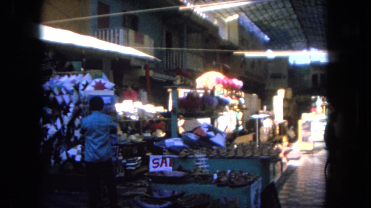 1962 Tijuana market interior with a shopper in a light shirt, display counters, sale signage, tiled floor, and bright overhead lighting.